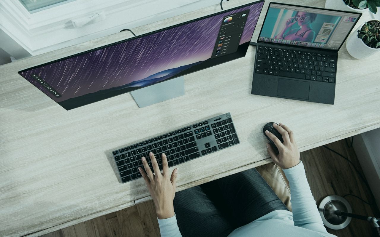 Designer working across a desktop monitor and laptop at a bright desk